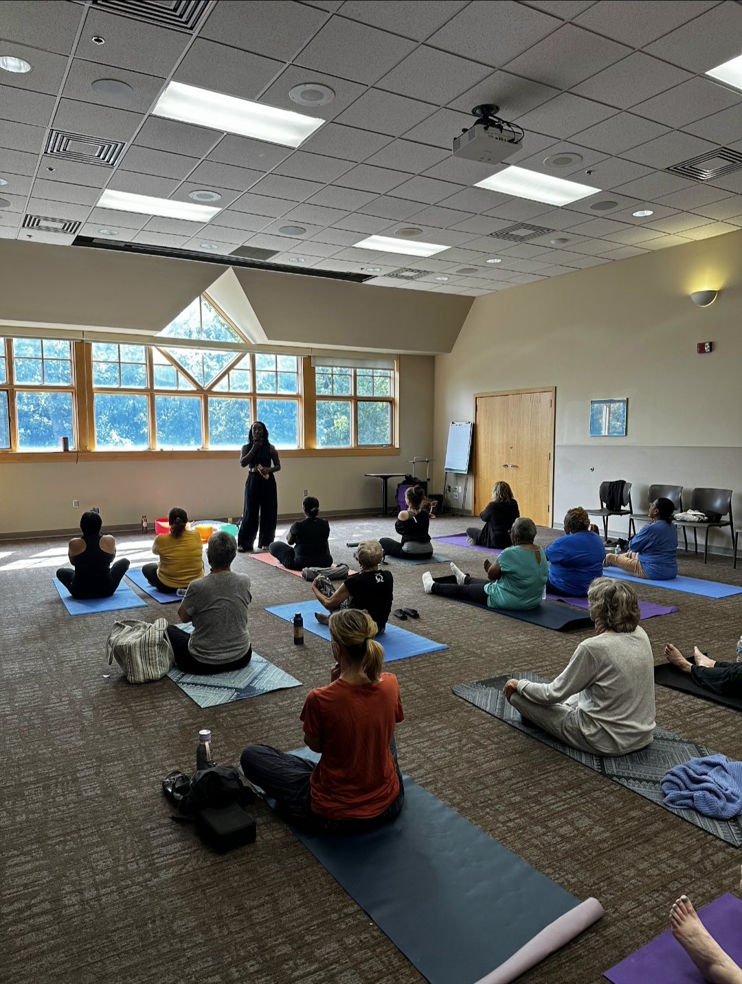 A seated meditation class in a carpeted room with tall windows, held from the front of the room