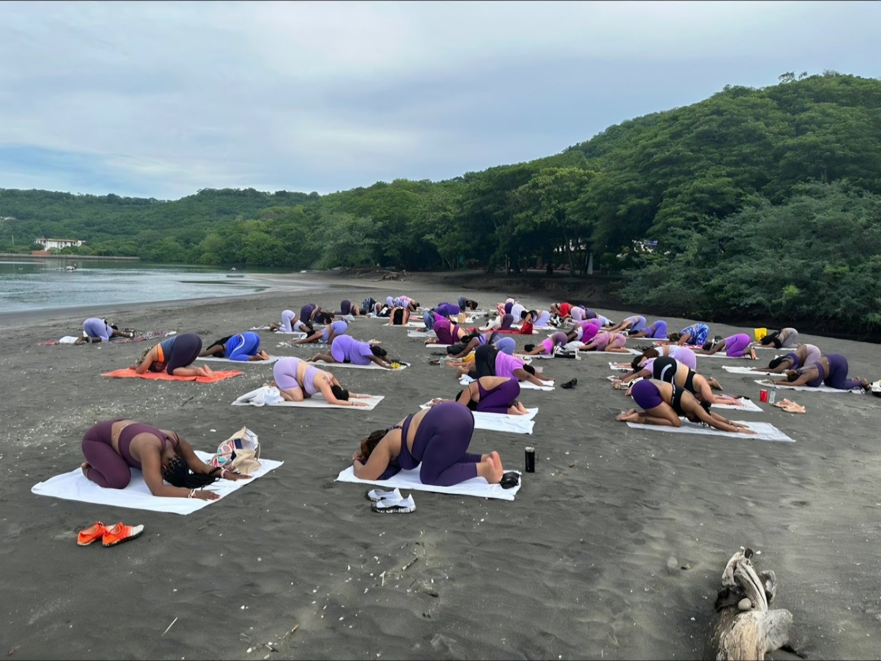 A group moving through forward folds on towels along a dark-sand beach backed by green hills
