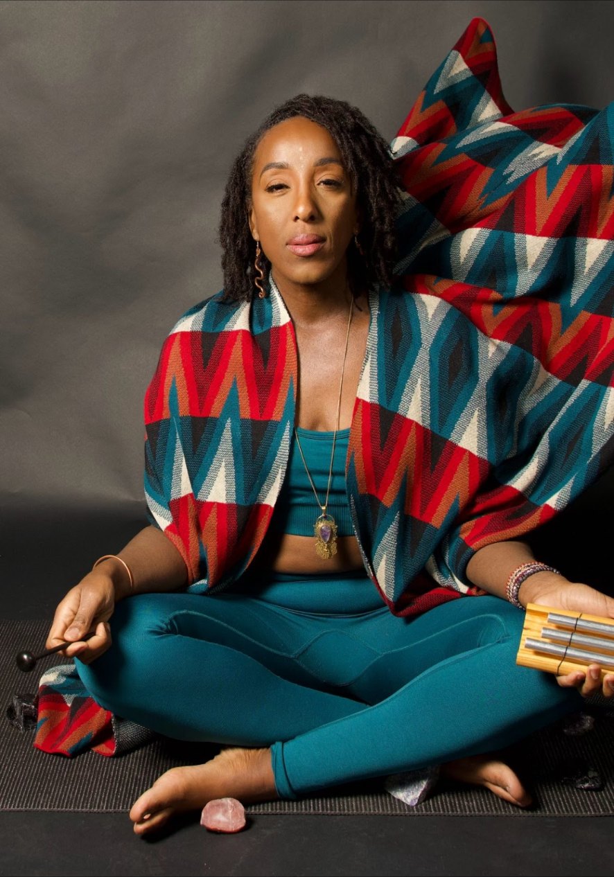 Andrea seated cross-legged on a mat holding chime bars and a mallet, wrapped in a patterned shawl
